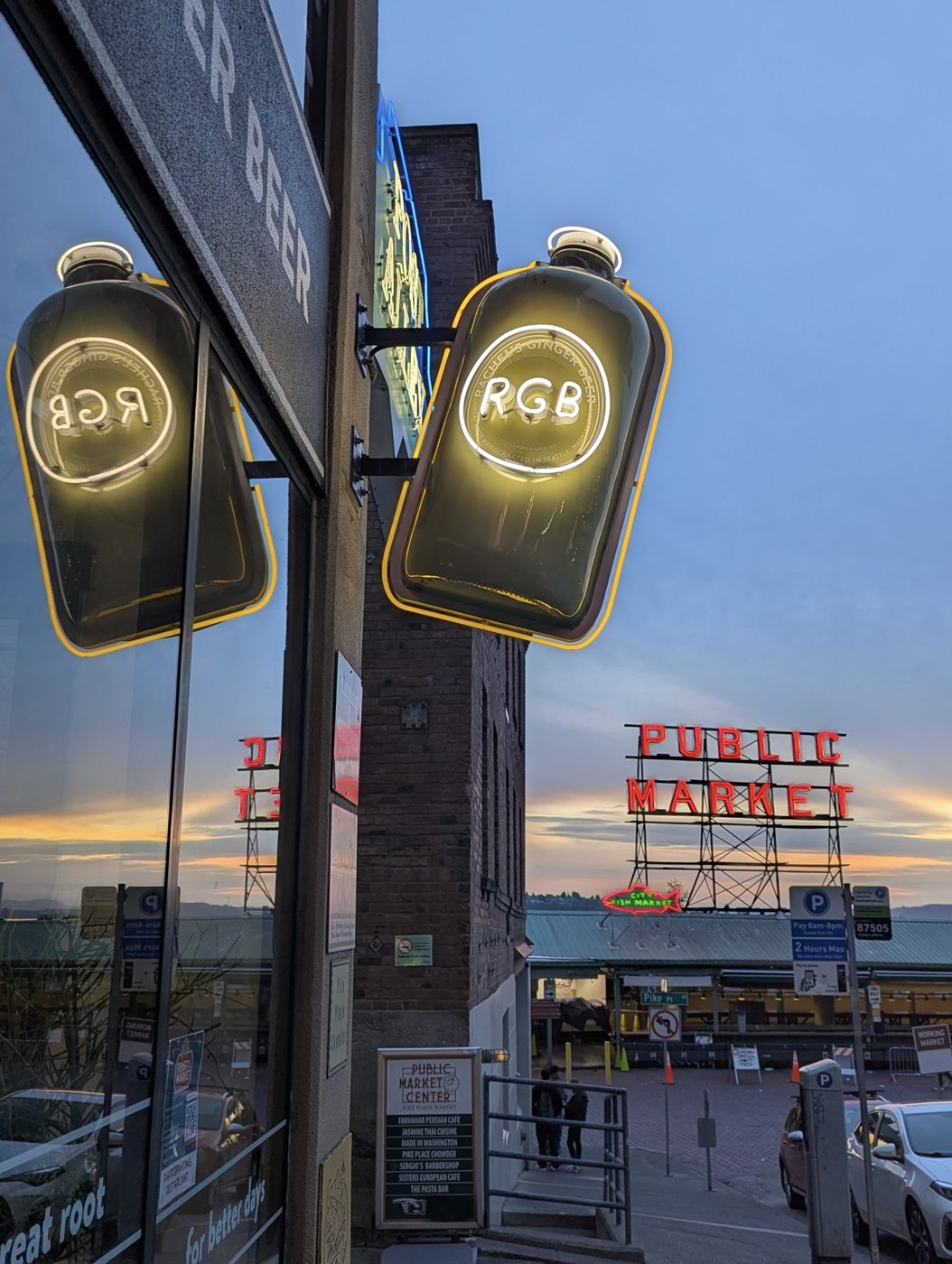 Neon signs at Pike Place Market at dusk: a glowing ginger beer bottle sign and the red Public Market sign against a sunset sky